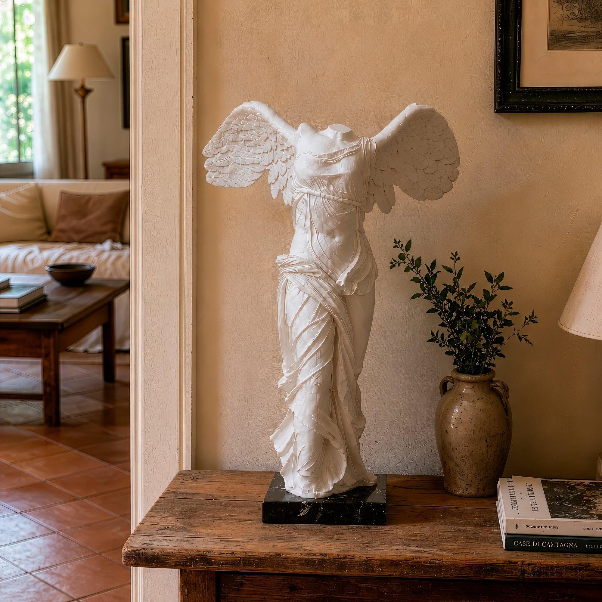 Reproduction of the Nike of Samothrace, a classical white marble statue depicting a winged female figure without head and arms, with flowing drapery and outstretched wings, on a dark base, viewed from the front