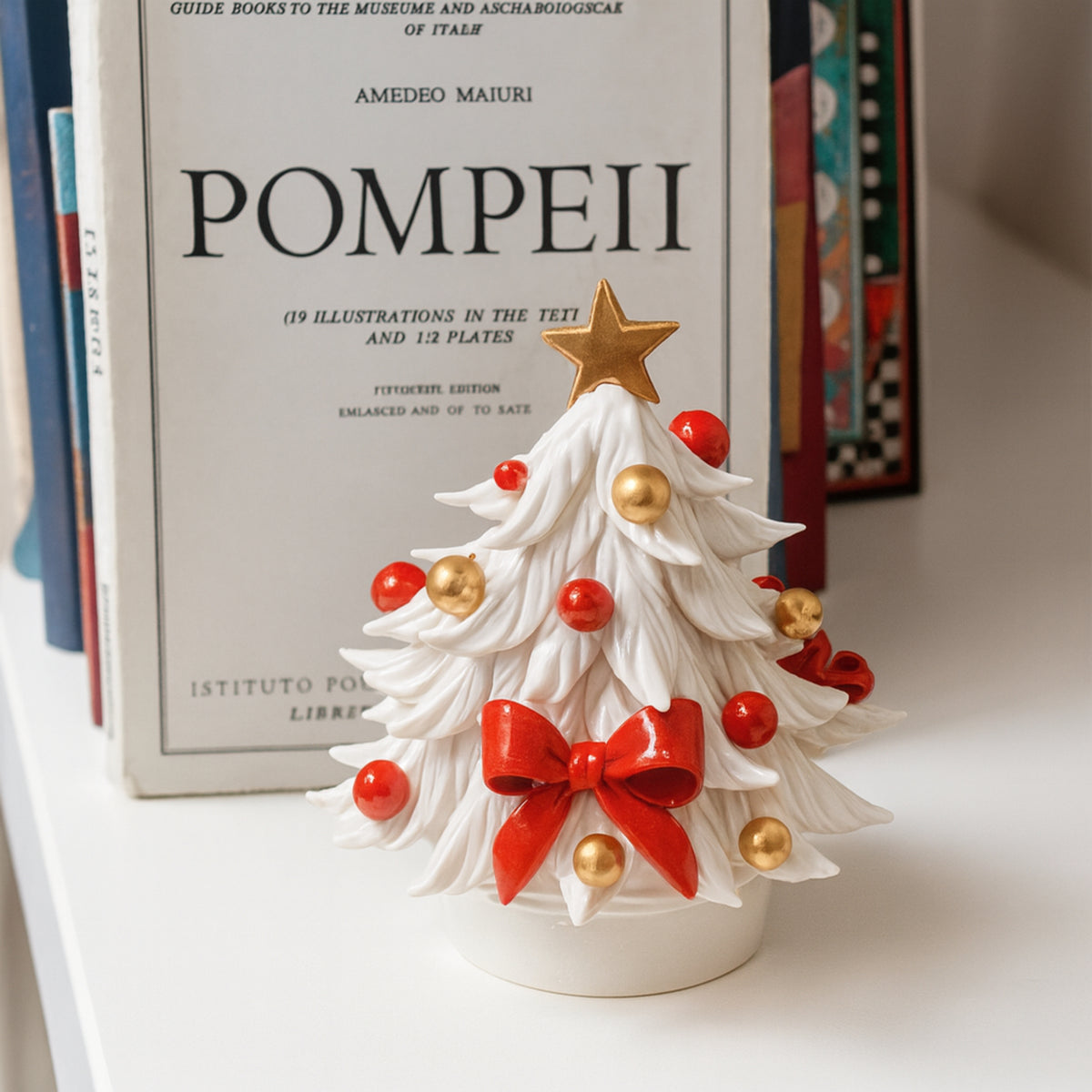 Albero di Natale bianco in porcellana con fiocco rosso, palline rosse e dorate e stella dorata, davanti ai libri