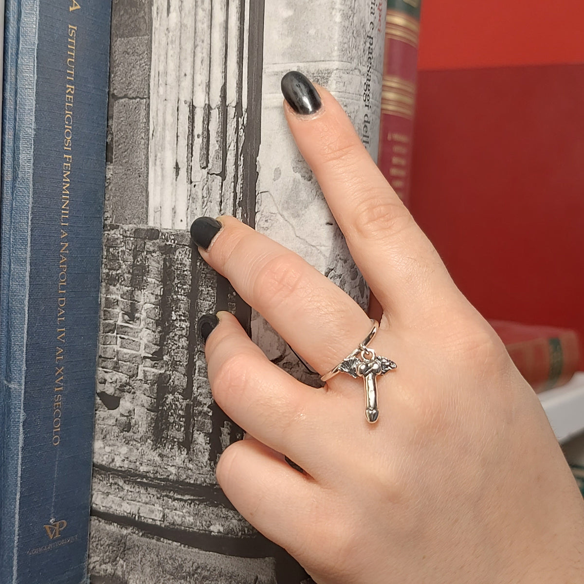 Anello in argento con pendente a fallo alato, indossato sulla mano appoggiata a libri