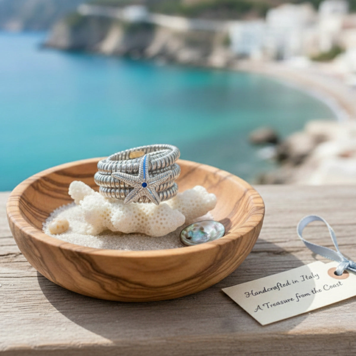 Anello con stella marina e centro blu, in una ciotola di legno con sabbia e conchiglie, con il mare sullo sfondo