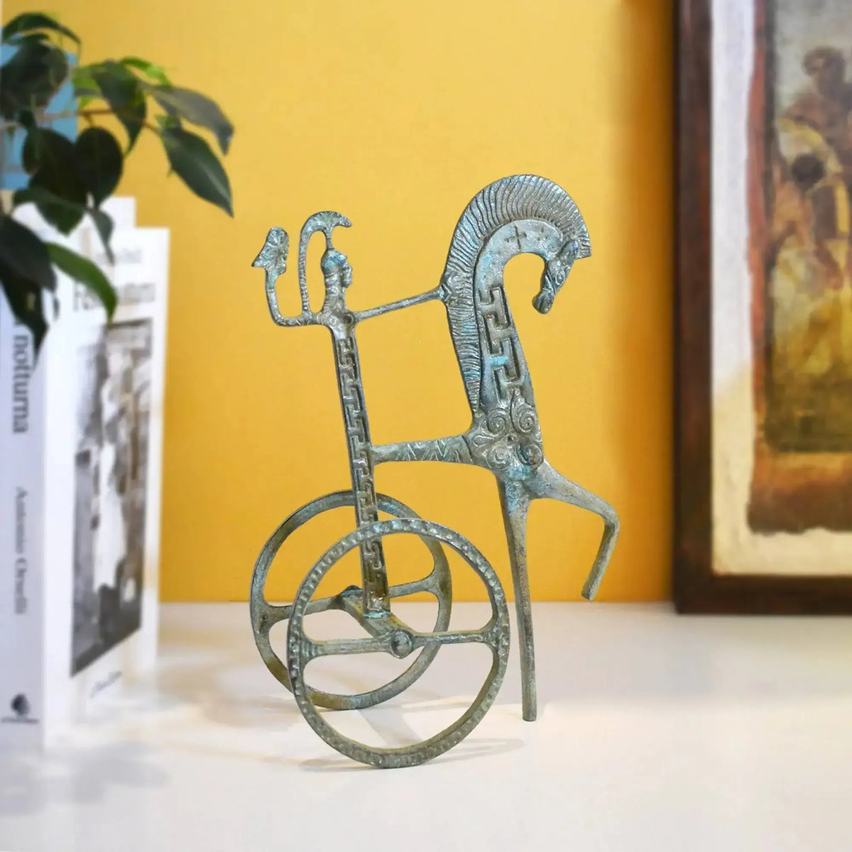 Biga greca in bronzo con patina verde su un piano bianco, accanto a libri e un quadro