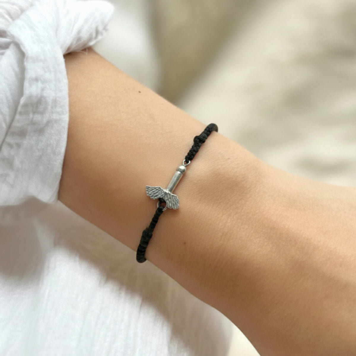 Bracciale in cotone nero con fallo alato in argento, indossato al polso accanto a una camicia bianca