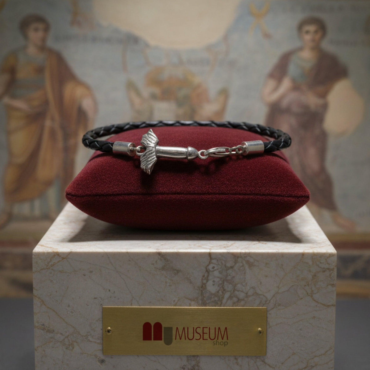 Bracciale in filo nero intrecciato con fallo alato in argento, su un cuscino rosso davanti a una parete dipinta
