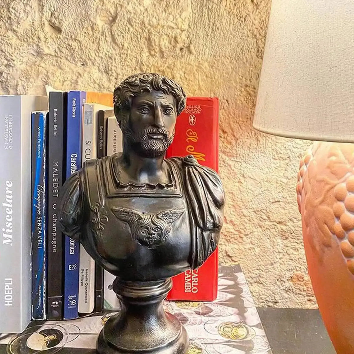 Busto di Adriano imperatore romano in bronzo scuro, con barba, armatura decorata e base rotonda, fotografato accanto a libri e lampada.