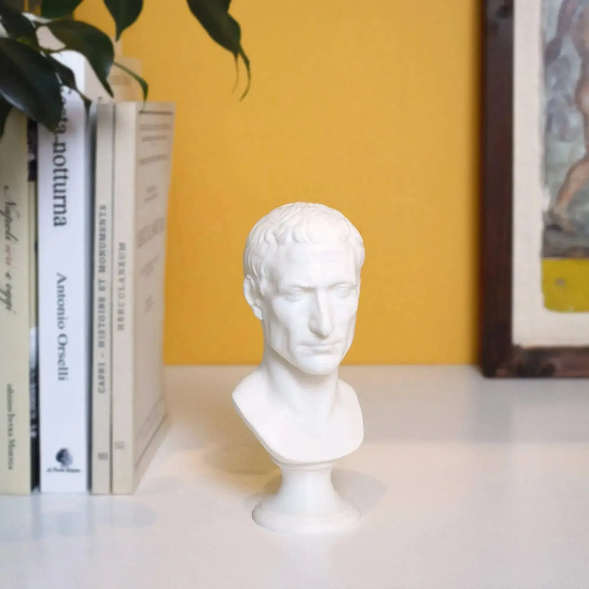Busto di Giulio Cesare stampato in 3D bianco su una mensola accanto a libri e parete gialla.