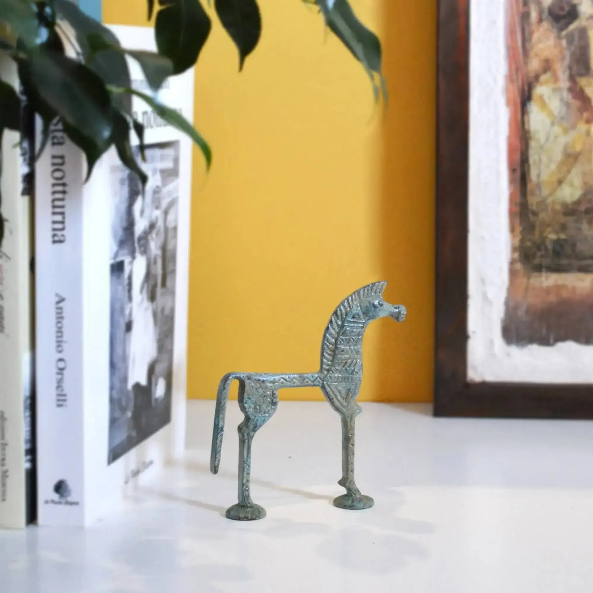 Cavallo greco in bronzo con patina verde-azzurra, forma stilizzata e decorazioni in rilievo, fotografato in ambiente con libri e quadro sullo sfondo.