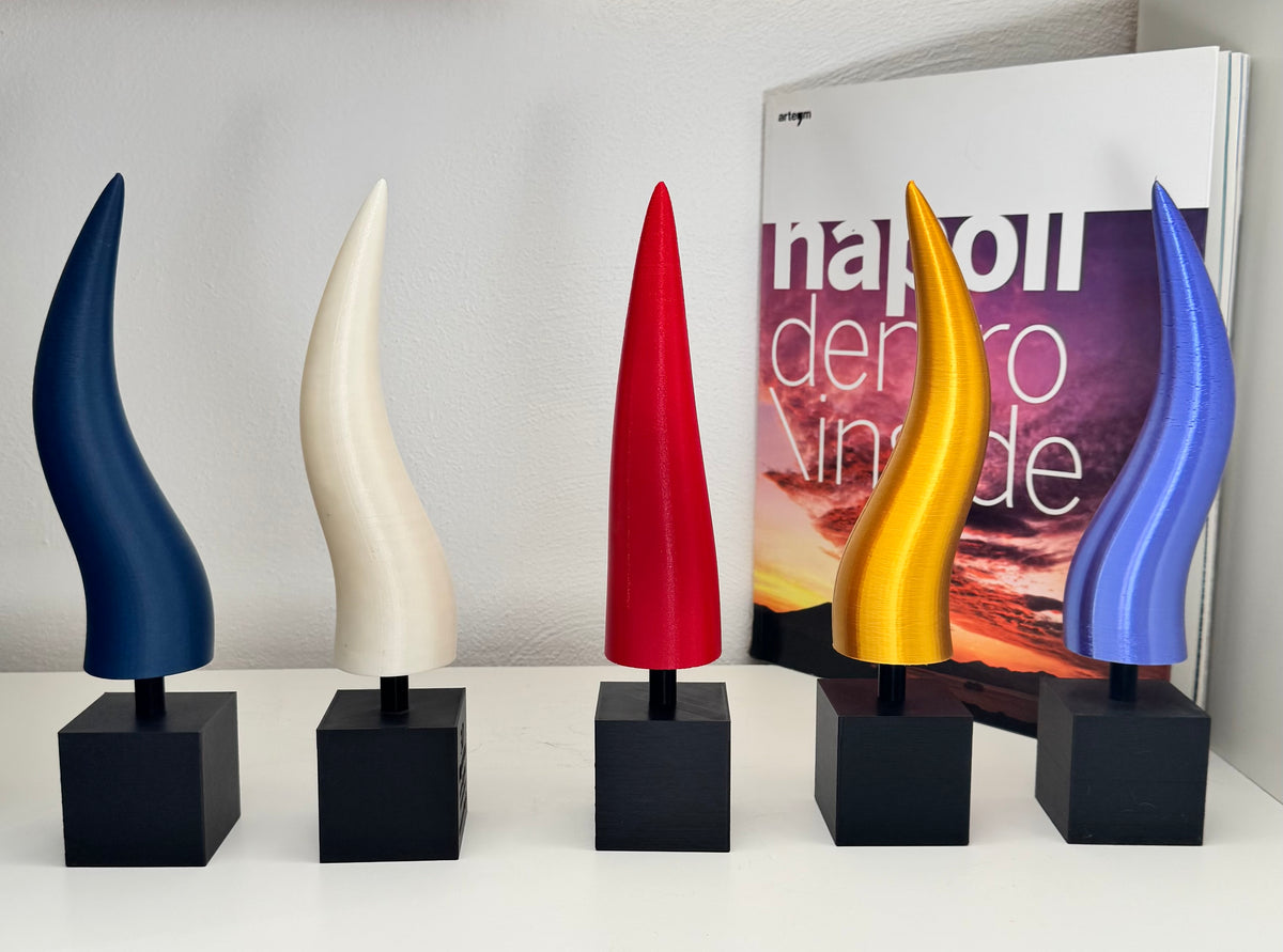 Cinque corni portafortuna in PLA di colori diversi su basi nere, esposti su una mensola davanti a un libro.