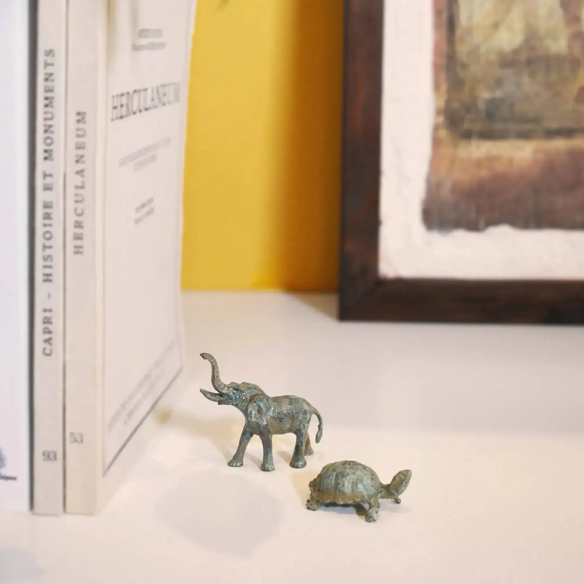Elefante in bronzo con proboscide alzata, accanto a una tartaruga su un ripiano con libri e un quadro
