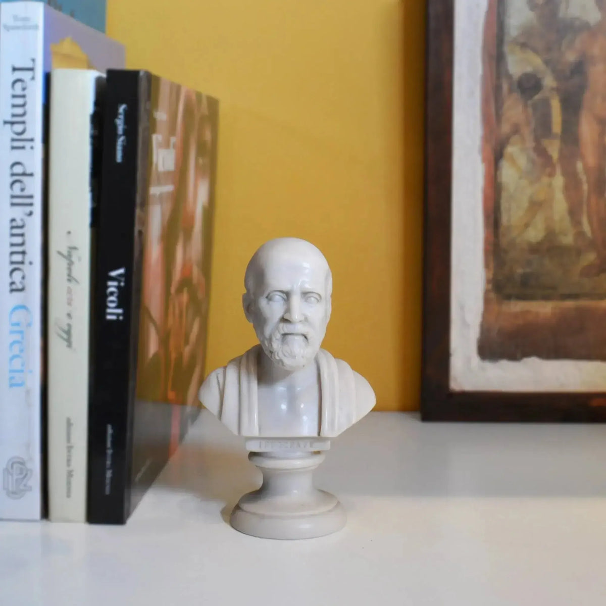 Busto di Ippocrate in marmo ricomposto su una mensola accanto a libri, con parete gialla e quadro sullo sfondo.