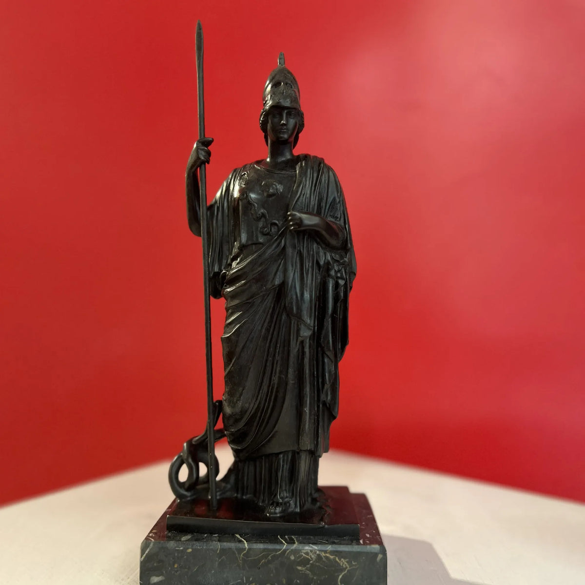 Minerva in bronzo scuro con elmo e lancia, vista frontale su base in marmo contro una parete rossa.