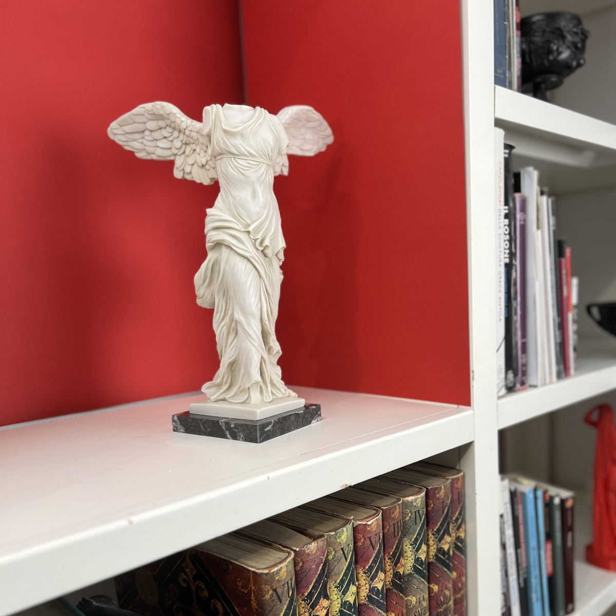 Nike di Samotracia in marmo ricomposto su un ripiano di una libreria, davanti a una parete rossa