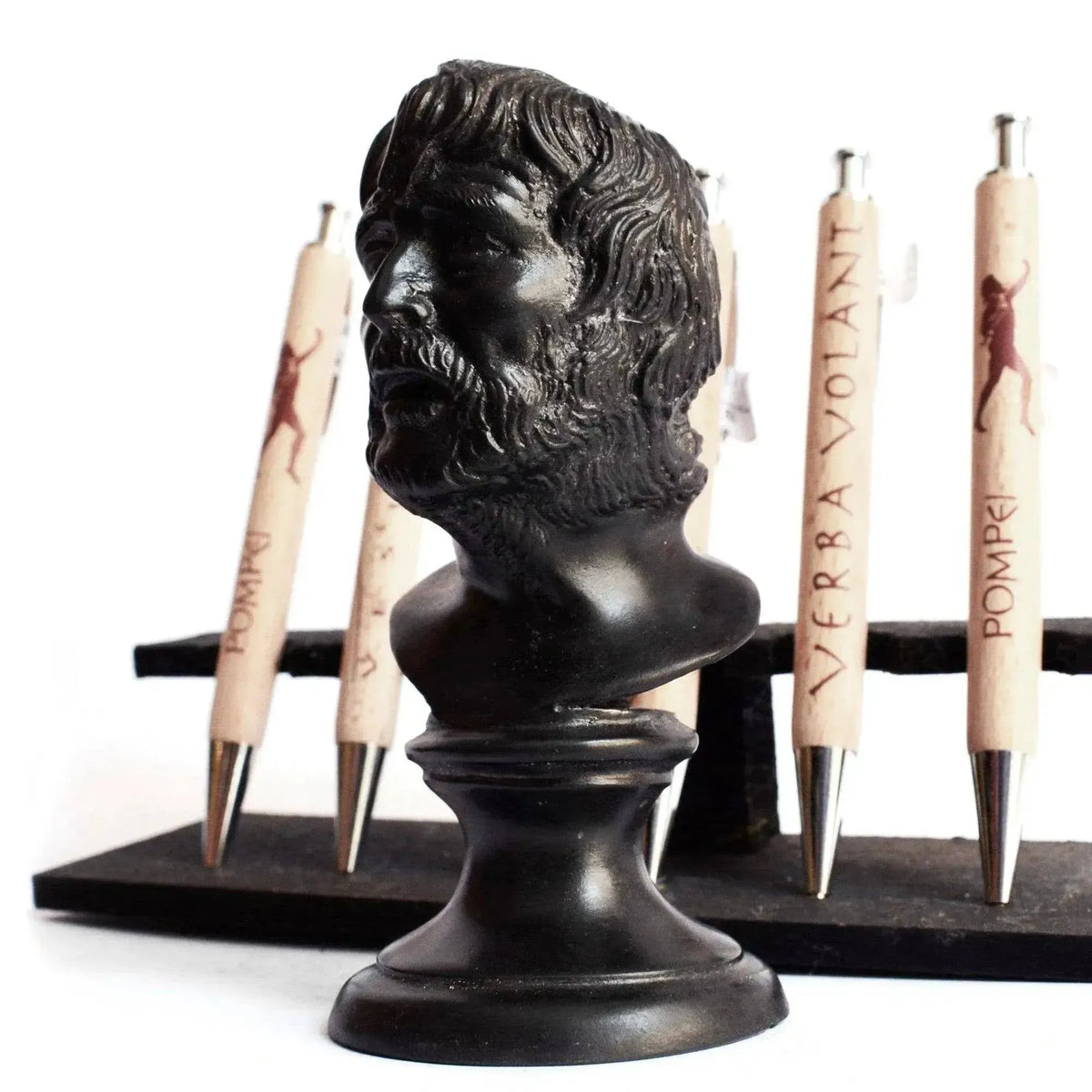 Testa di Seneca in bronzo scuro su base rotonda, con dettagli scolpiti di barba e capelli, fotografata di profilo con penne decorative sullo sfondo.