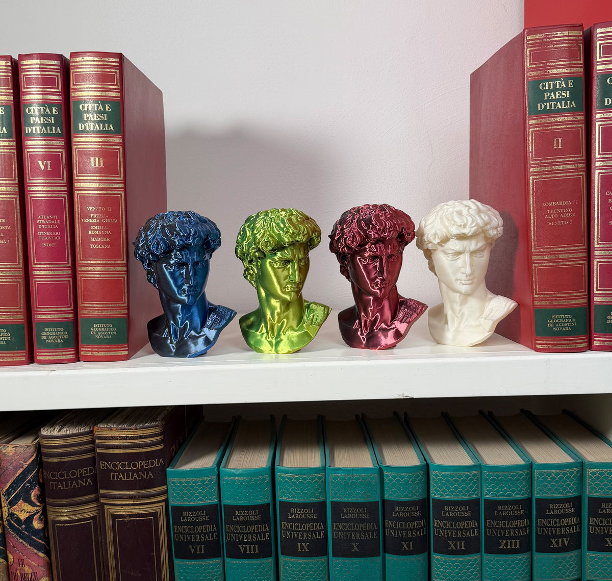 Quattro teste del David in PLA di colori diversi, viste frontalmente su una mensola tra libri.