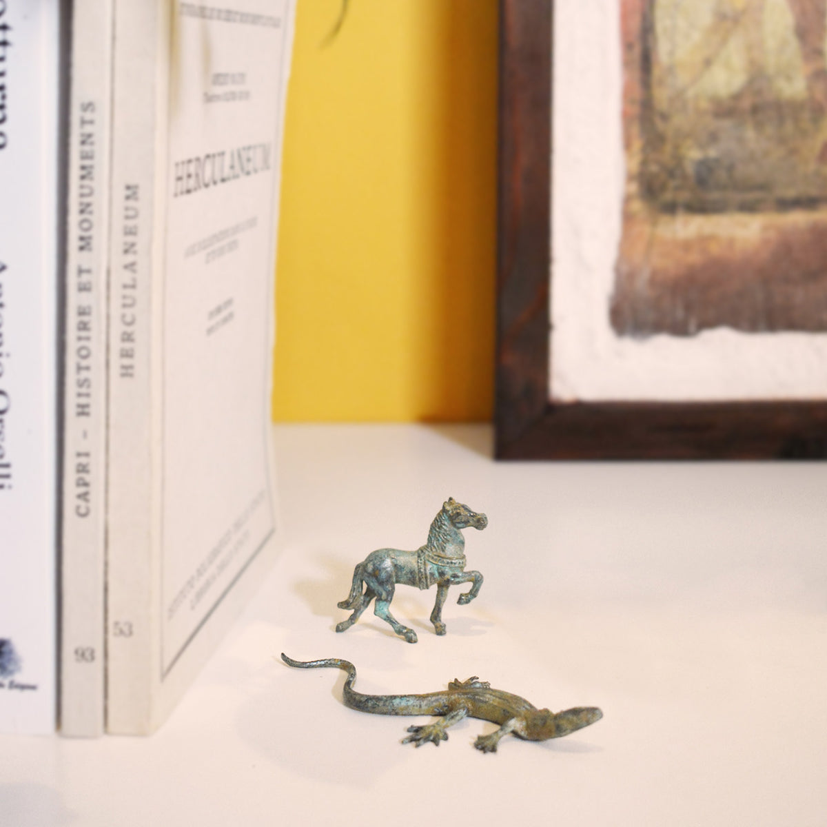 Cavallo greco mignon e lucertola in bronzo su un piano bianco, accanto a libri