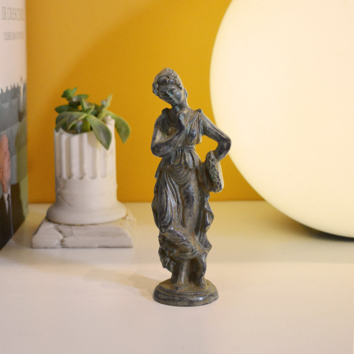 Roman woman in bronze with green patina, draped dress and decorative base, photographed in a setting with a plant and warm light in the background.