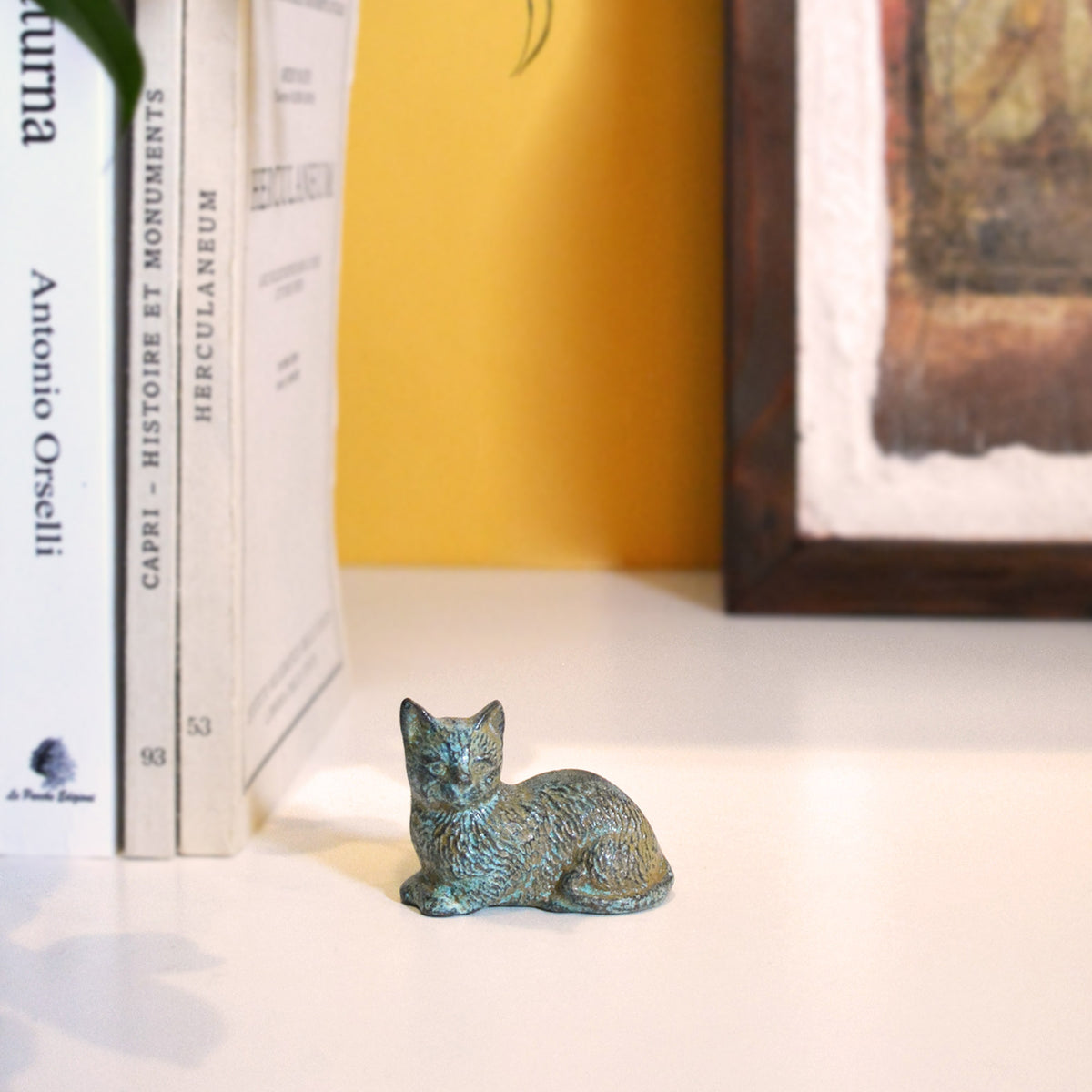 Gatto accovacciato in bronzo su un ripiano bianco, accanto a libri e un quadro