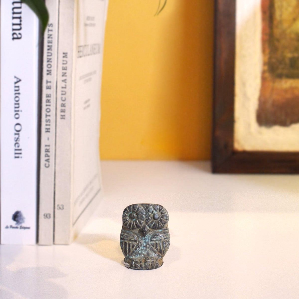 Gufo in bronzo con patina verde su una mensola accanto a libri, con parete gialla sullo sfondo.