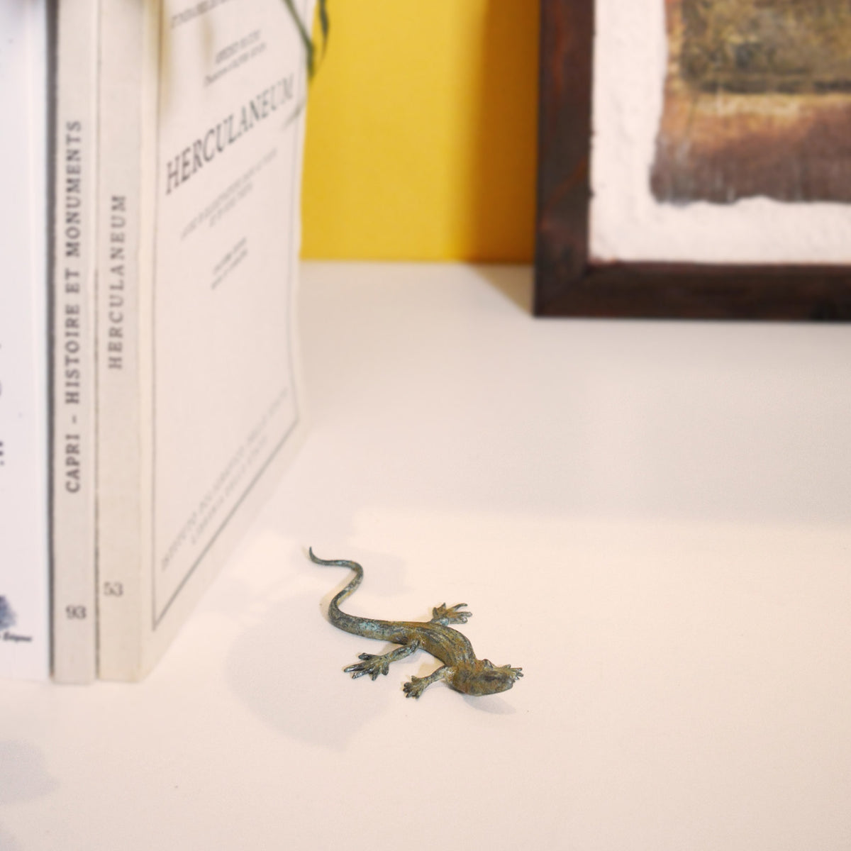 Lucertola in bronzo con patina verde su una mensola accanto a libri, con parete gialla e quadro sullo sfondo.