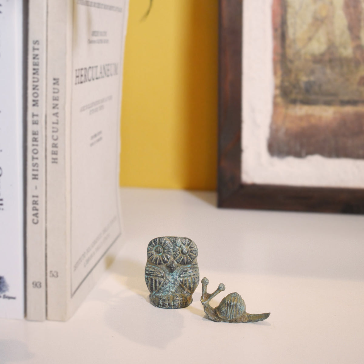 Lumaca in bronzo con patina verde accanto a un gufo decorativo, su una mensola con libri e parete gialla sullo sfondo.