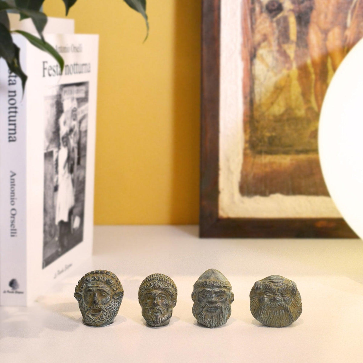 Quattro mini maschere etrusche in bronzo su una mensola accanto a libri, con parete gialla e quadro sullo sfondo.