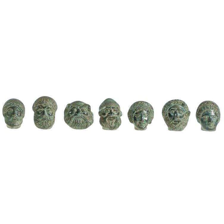 Sette mini maschere etrusche in bronzo con patina verde, disposte in fila su fondo bianco.